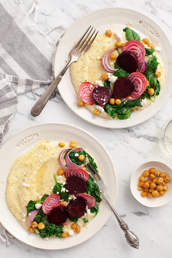 Two plates of creamy polenta topped with sautéed greens, sliced beets, chickpeas, and feta cheese, placed on a marble surface with a striped napkin and a fork.