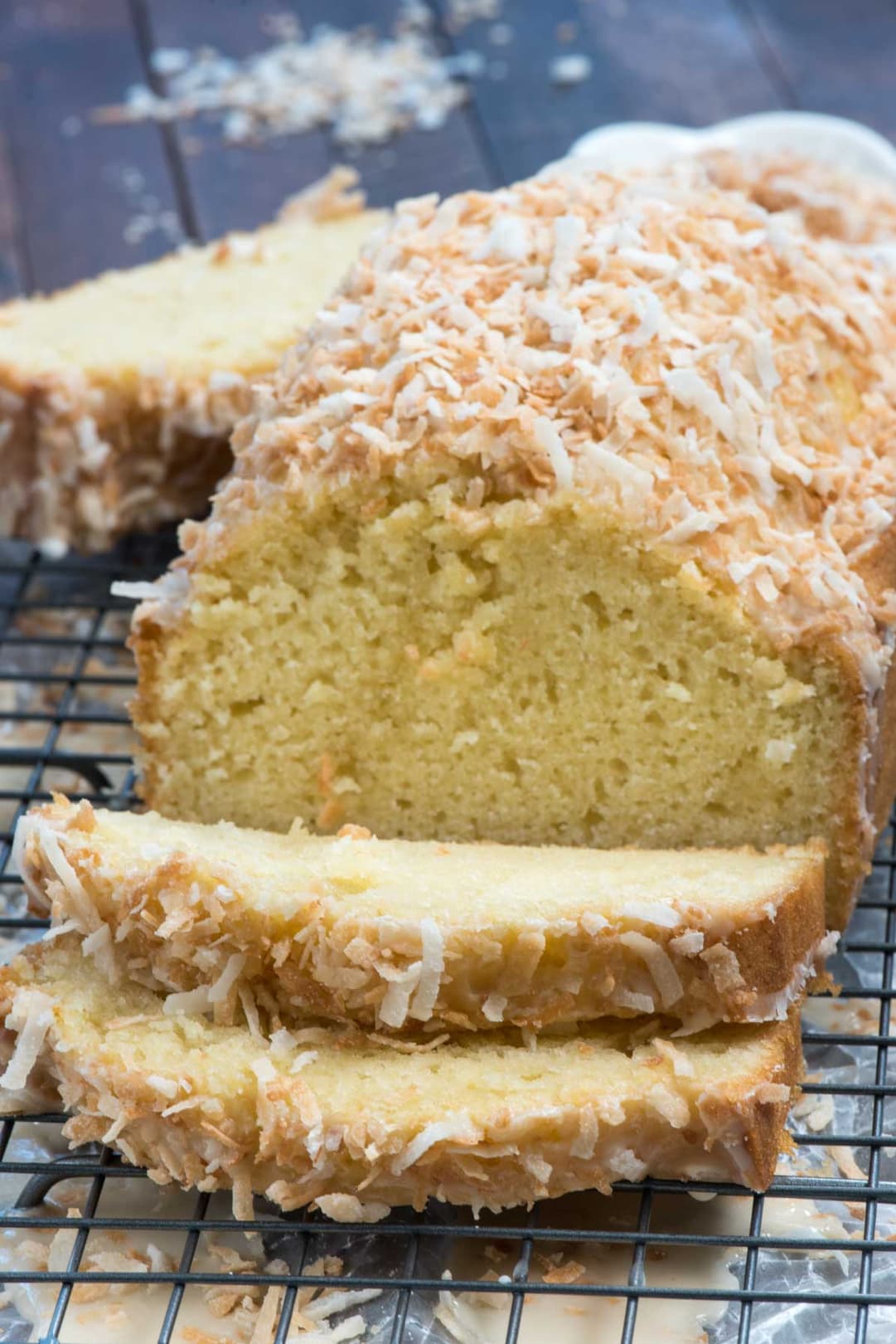 A loaf of coconut bread topped with shredded coconut is sliced and placed on a cooling rack, with crumbs and coconut scattered around.