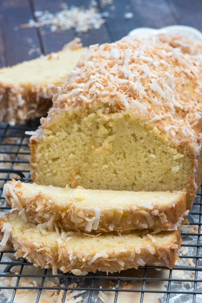 A loaf of coconut bread topped with shredded coconut is sliced and placed on a cooling rack, with crumbs and coconut scattered around.