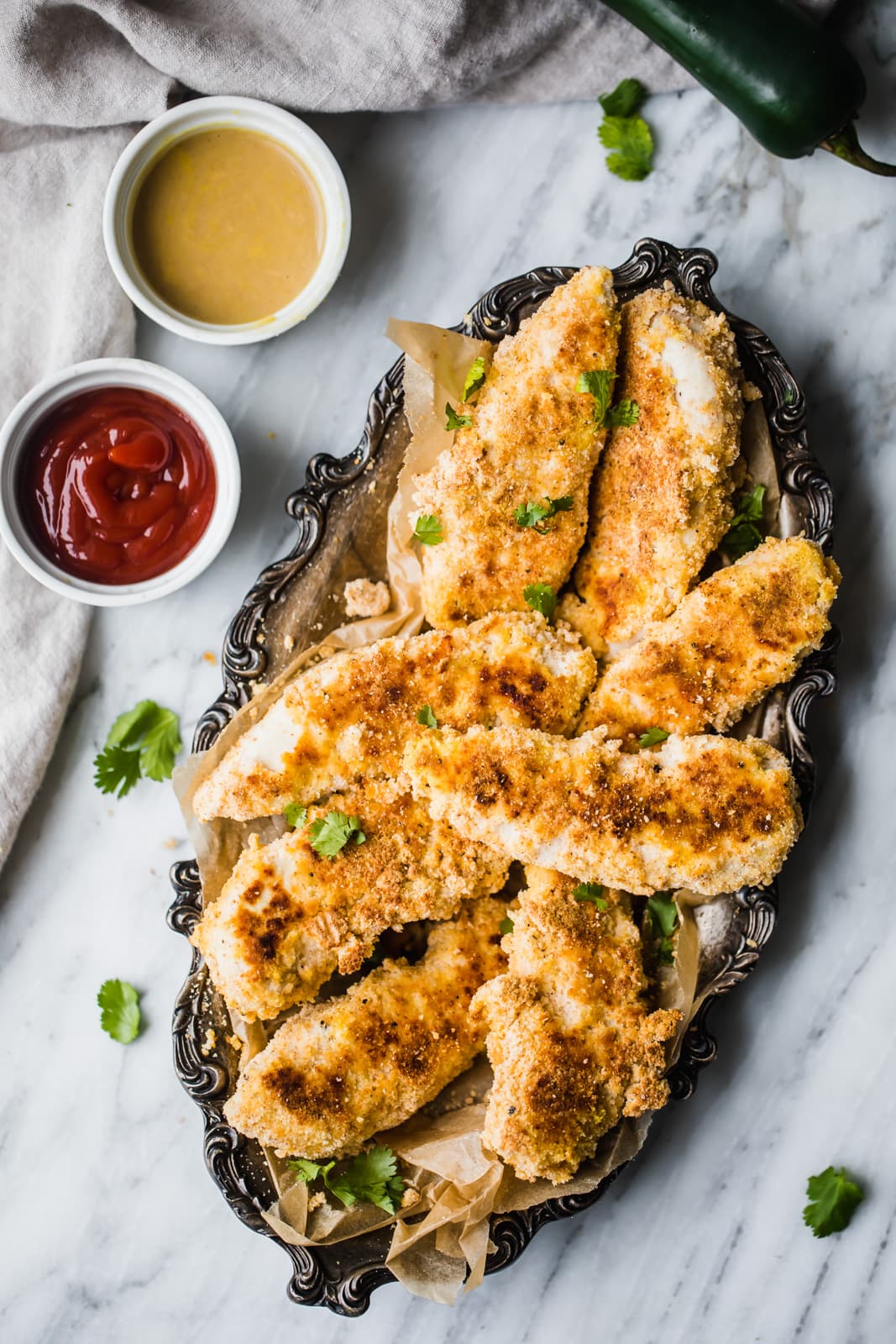 A tray of breaded chicken tenders garnished with herbs, served with ketchup and honey mustard dipping sauces on a marble surface.