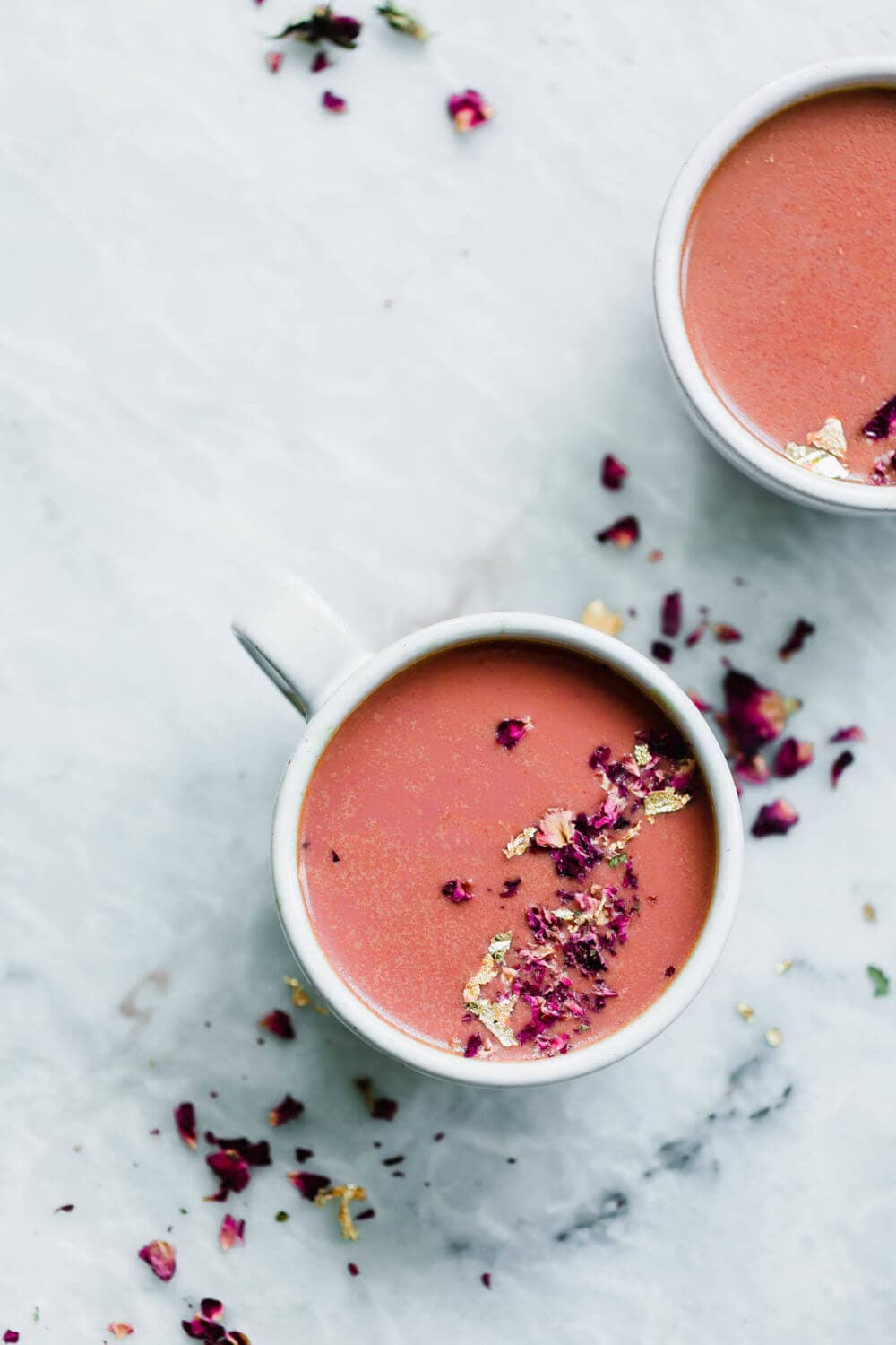 A white cup filled with pink rose latte topped with dried rose petals and gold leaf sits on a light marble surface, next to another partially visible cup.