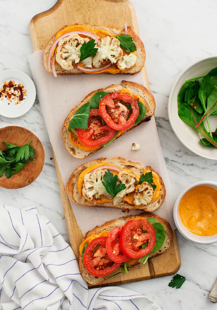 Three open-faced sandwiches with roasted cauliflower, tomatoes, spinach, and sauce on a wooden board, surrounded by fresh greens, parsley, and a small bowl of sauce.