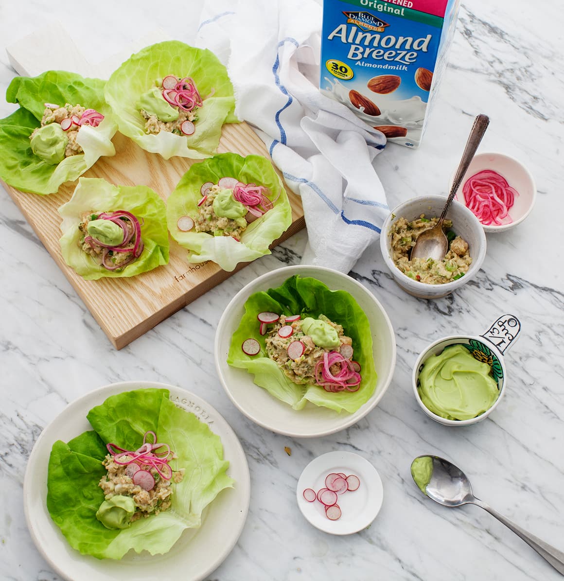 Lettuce wraps filled with a chickpea mixture, avocado sauce, and pickled onions on a marble table with a carton of almond milk and sliced radishes.