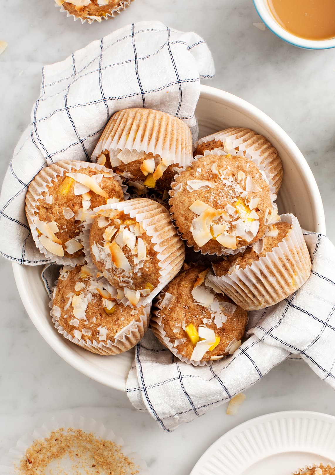 A bowl lined with a checkered cloth holds several muffins topped with coconut flakes, next to a cup of coffee and a plate on a marble surface.