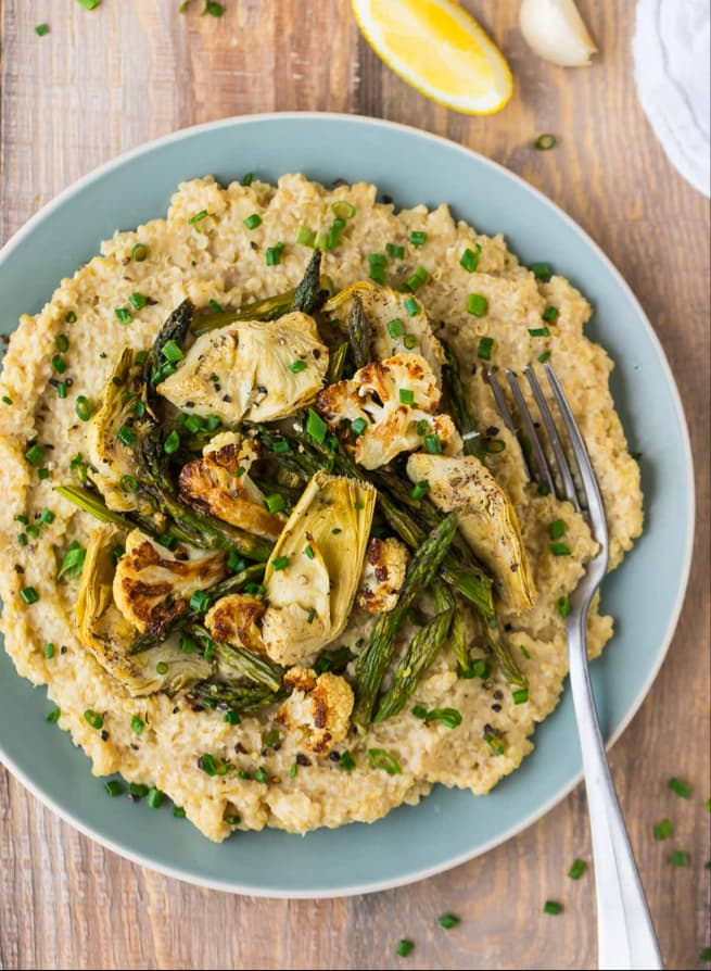 A plate of creamy risotto topped with roasted asparagus, cauliflower, and artichoke hearts, garnished with chopped chives. A fork rests on the plate; lemon wedges are nearby.