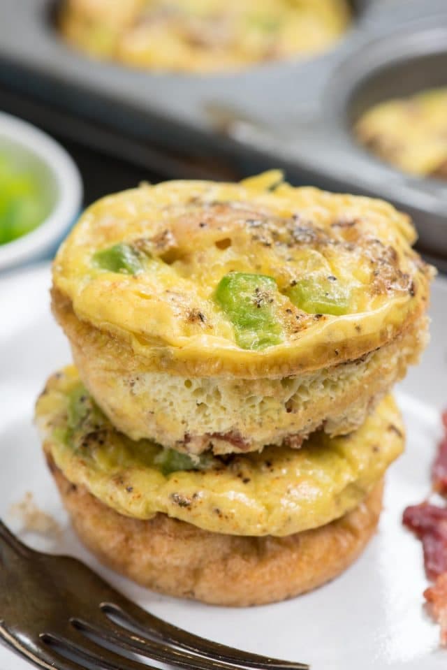 Two egg muffins with green bell pepper pieces are stacked on a white plate next to a fork and a strip of bacon, with a muffin tin visible in the background.