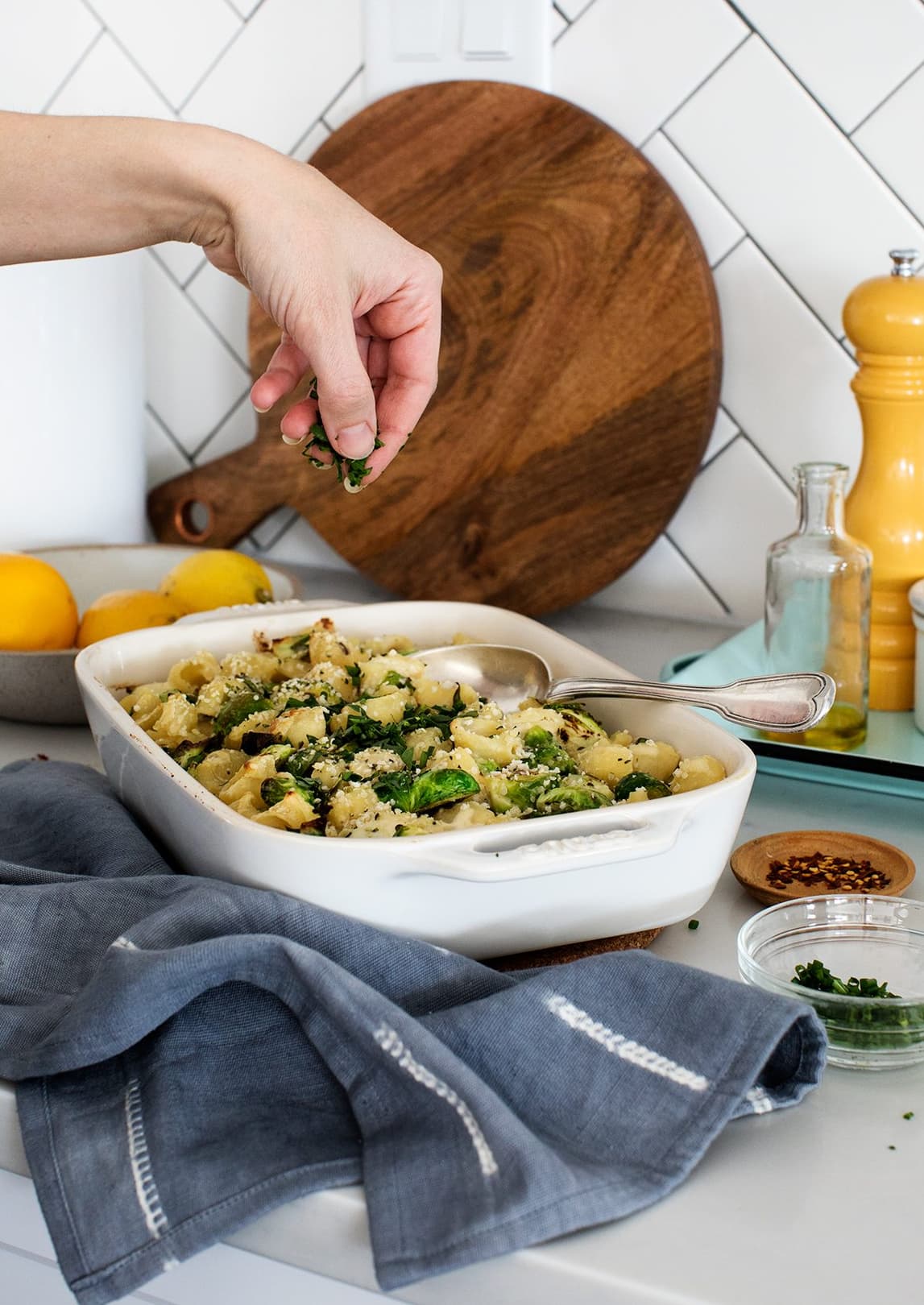 A hand sprinkles herbs over a baked pasta dish in a white casserole on a kitchen counter, with lemons, a cutting board, and seasonings in the background.