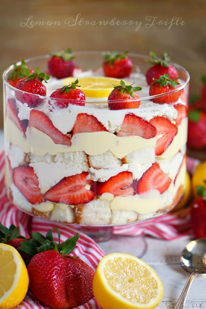 A glass trifle dish filled with layers of whipped cream, cake, custard, and sliced strawberries, topped with whole strawberries and lemon slices. Lemons and strawberries are beside the dish.