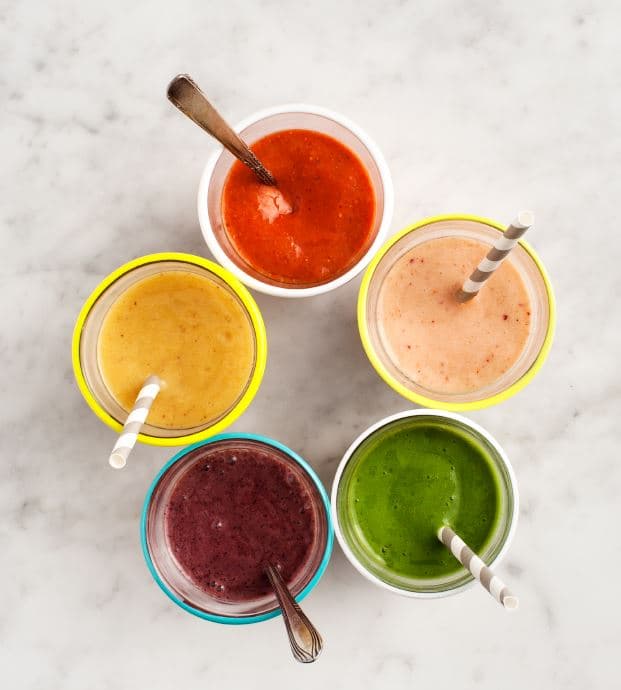 Five cups of smoothies in different colors—red, yellow, purple, green, and beige—on a white marble surface. Each cup has a straw or spoon.