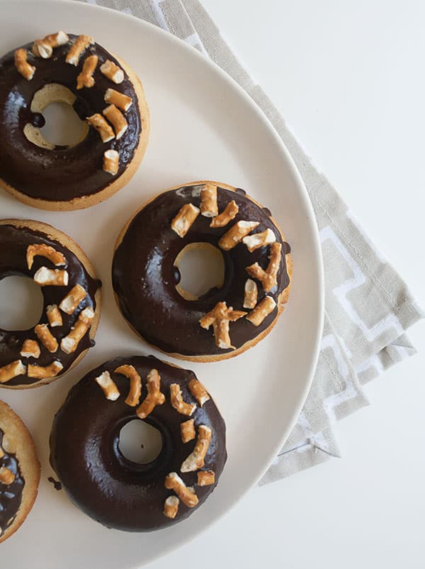 Four donuts with chocolate glaze and pretzel pieces on a white plate, set on a light-colored patterned napkin.
