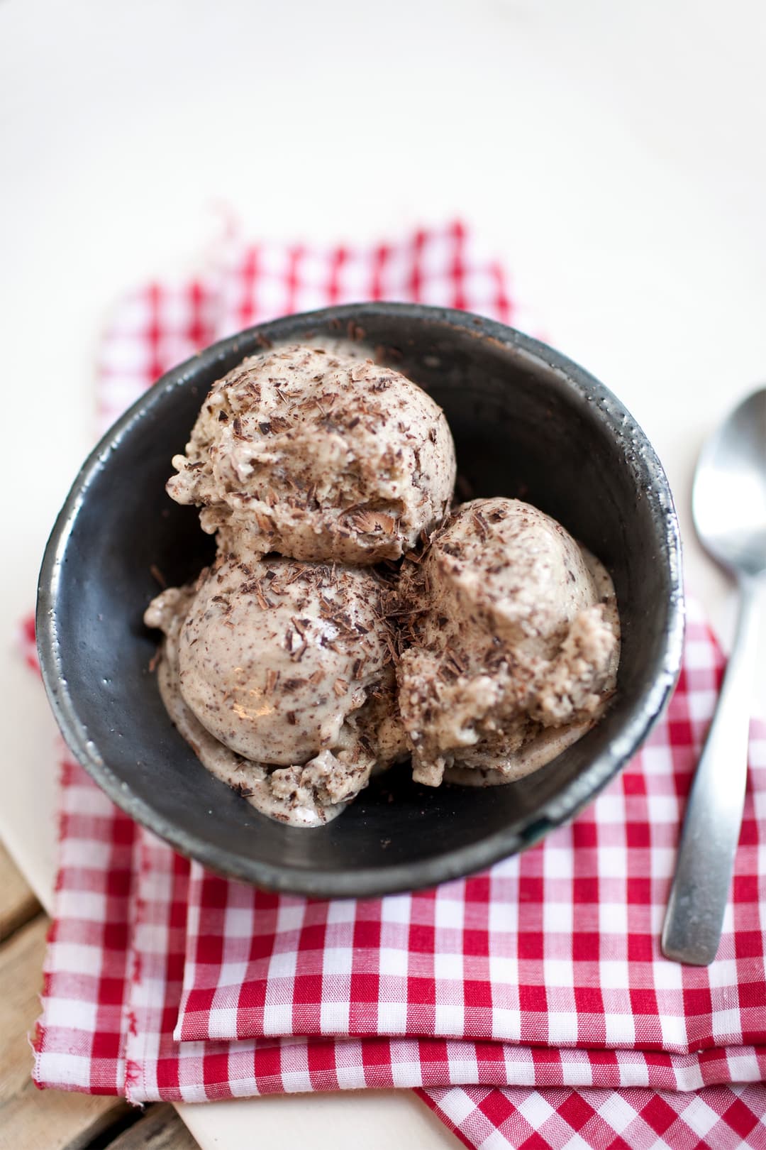 Three scoops of ice cream with chocolate shavings in a dark bowl, placed on a red and white checkered cloth next to a metal spoon.