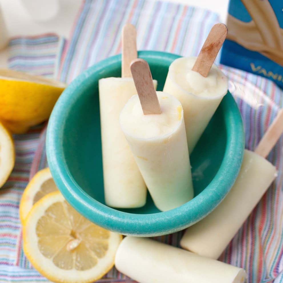Four creamy popsicles in a turquoise bowl, surrounded by lemon slices on a striped cloth, with a container labeled vanilla partially visible in the corner.