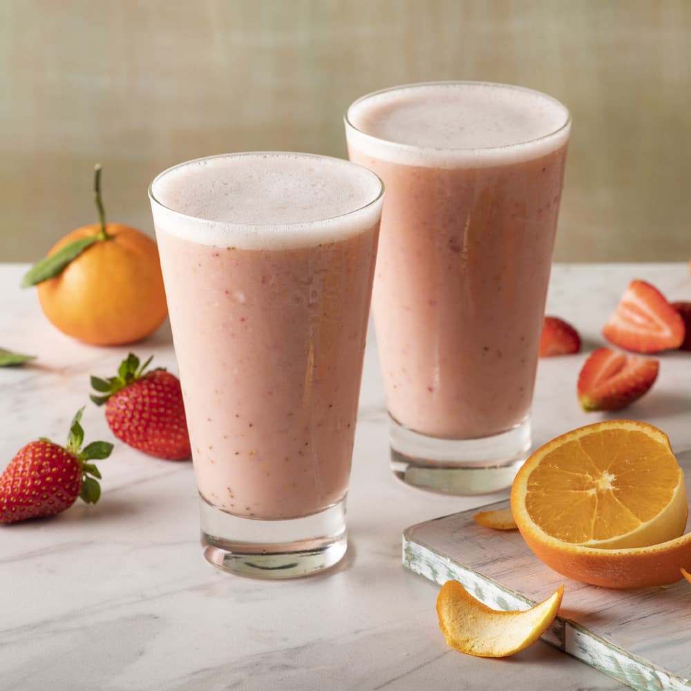 Two glasses of pink fruit smoothie on a marble surface, surrounded by strawberries, an orange cut in half, and a whole tangerine.