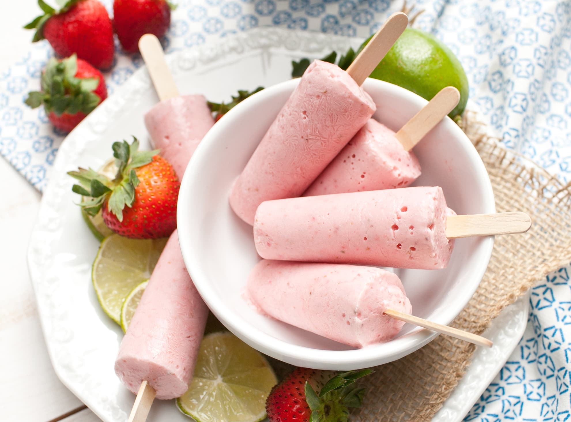 Five pink popsicles in a white bowl with fresh strawberries and lime slices on a plate, set on a textured placemat and patterned fabric.