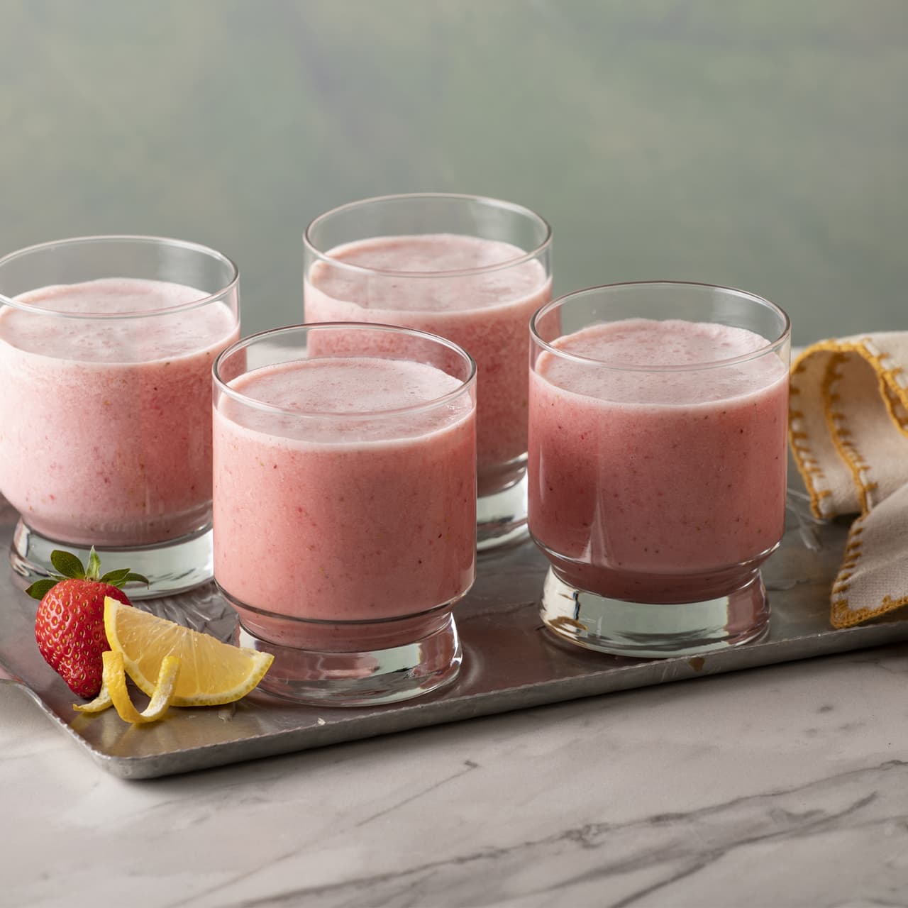 Four glasses of pink strawberry smoothie on a metal tray with a strawberry, lemon wedges, and a folded napkin on a marble surface.