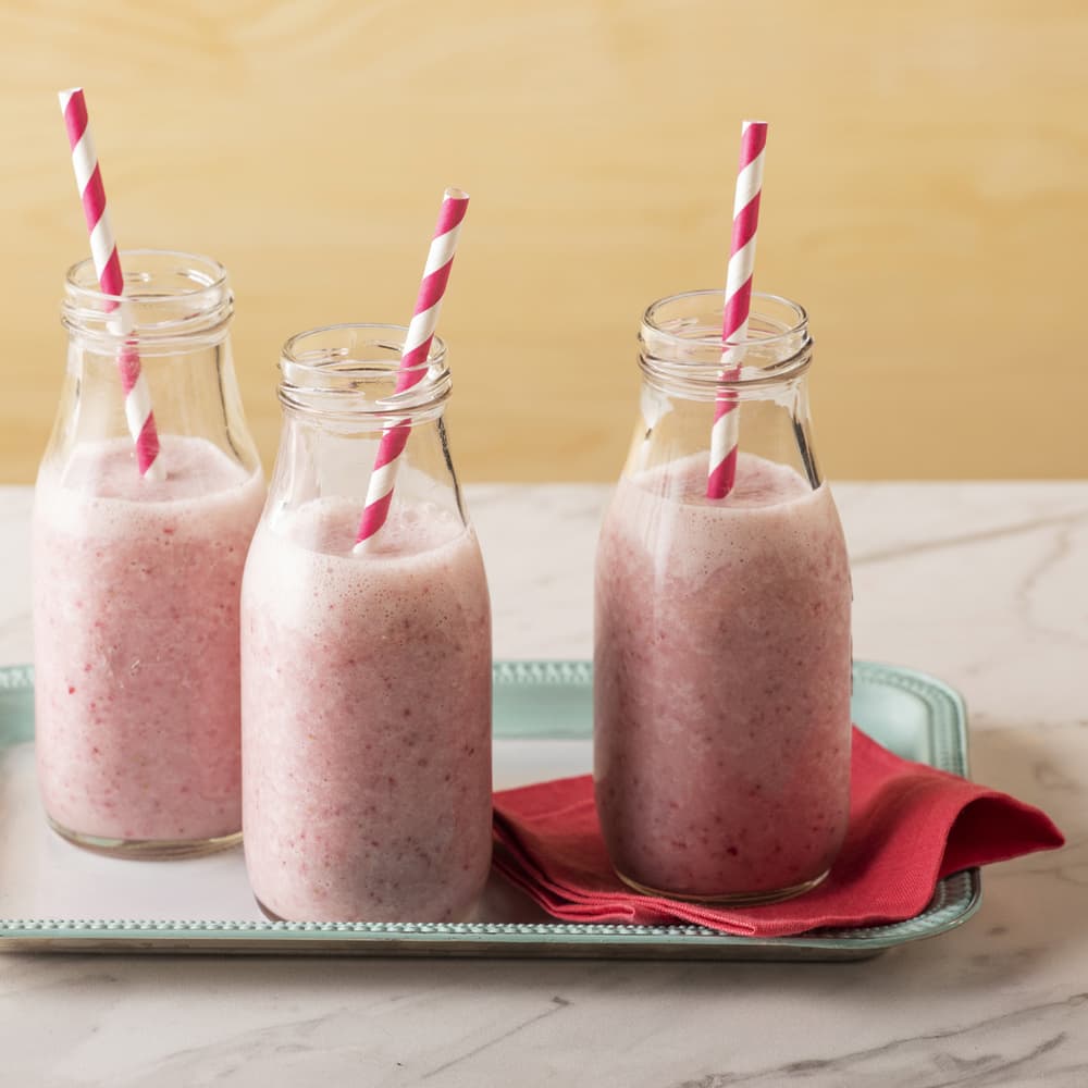 Three glass bottles filled with pink smoothies, each with a pink and white striped straw, are placed on a tray with a folded red napkin underneath.