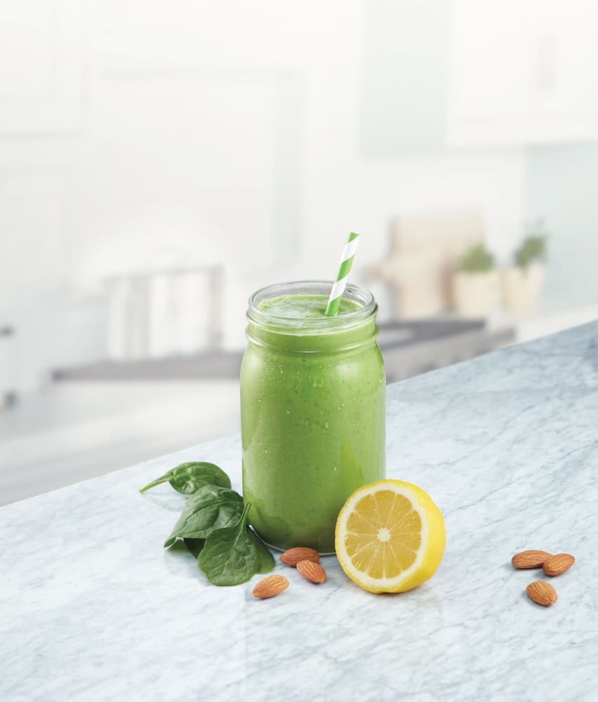 A green smoothie in a mason jar with a striped straw, next to spinach leaves, almonds, and a halved lemon on a marble counter.