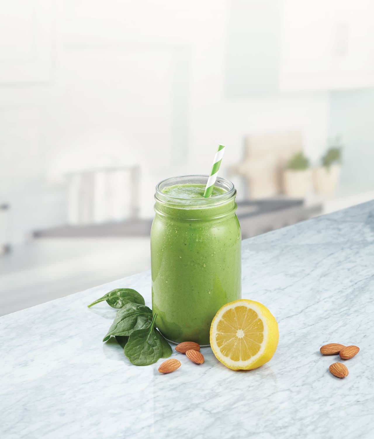 A green smoothie in a mason jar with a striped straw, next to spinach leaves, almonds, and a halved lemon on a marble counter.