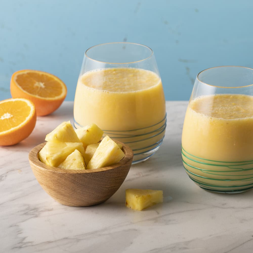 Two glasses of yellow smoothie on a marble surface, with a wooden bowl of pineapple chunks and a halved orange nearby, set against a light blue background.