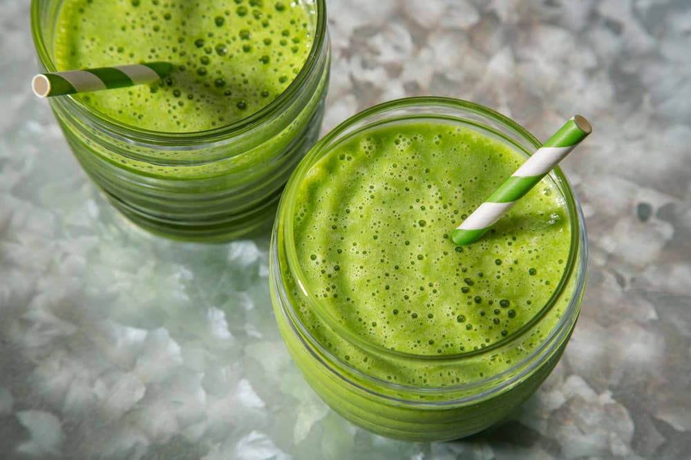 Two glass jars filled with green smoothies, each with a green and white striped paper straw, placed on a gray marbled surface.