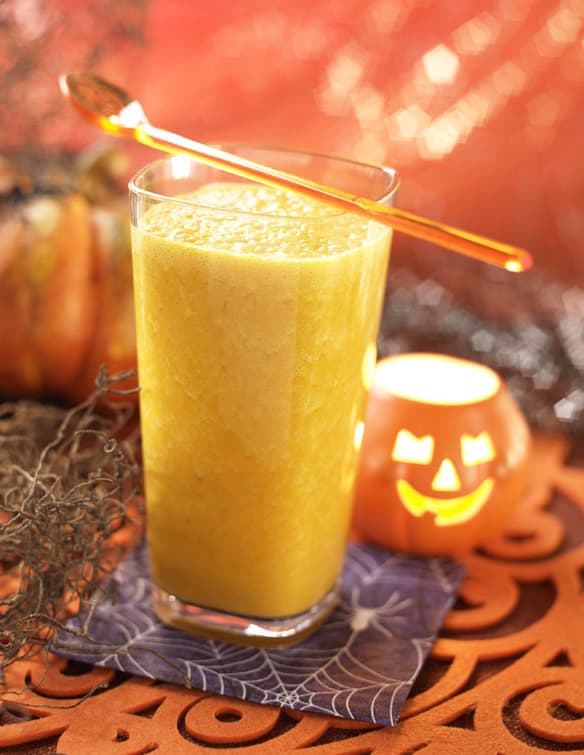 A glass of orange smoothie with a spoon on top sits on a Halloween-themed napkin, next to a jack-o-lantern candle and pumpkin, with festive decorations in the background.
