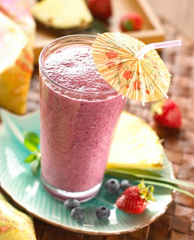 A glass of purple smoothie with a straw and paper umbrella sits on a turquoise plate, surrounded by pineapple slices, strawberries, and blueberries.