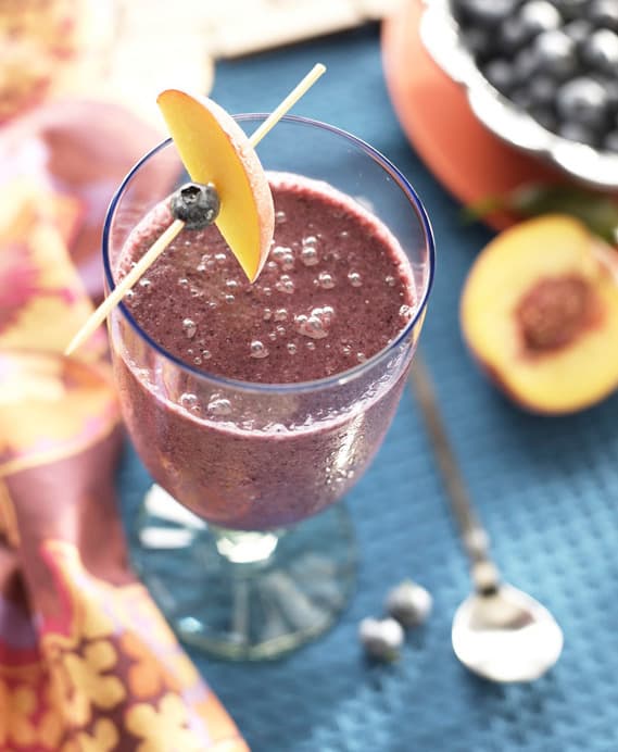 A glass of purple smoothie topped with a peach slice and blueberry garnish, set on a blue mat beside a spoon, blueberries, and a halved peach.