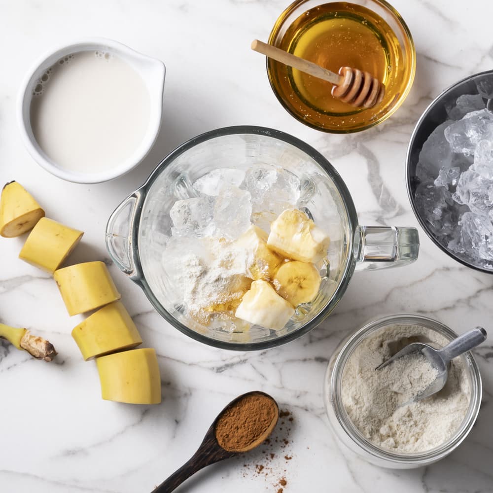 Overhead view of smoothie ingredients: banana, ice, milk, honey, sliced ginger, protein powder, and cinnamon on a marble surface.