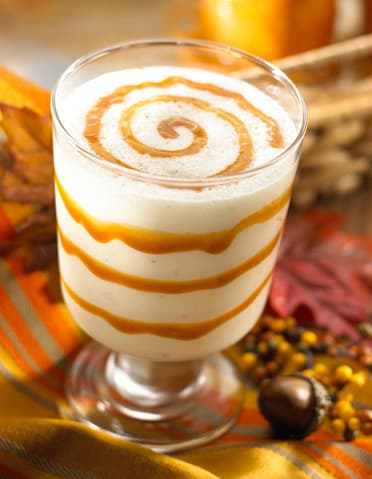 A glass filled with a creamy drink, topped and lined with swirls of caramel sauce, sits on a colorful autumn-themed cloth with decorative items nearby.