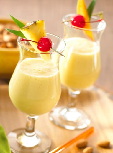 Two glasses of yellow tropical drink, each garnished with a pineapple slice, cherry, and a small green leaf, set on a wooden surface with nuts in the background.