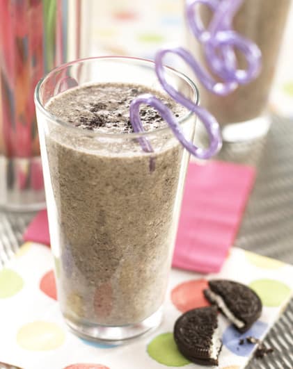 A glass of cookies and cream milkshake with a purple curly straw, placed on a polka-dot napkin next to a split chocolate sandwich cookie.