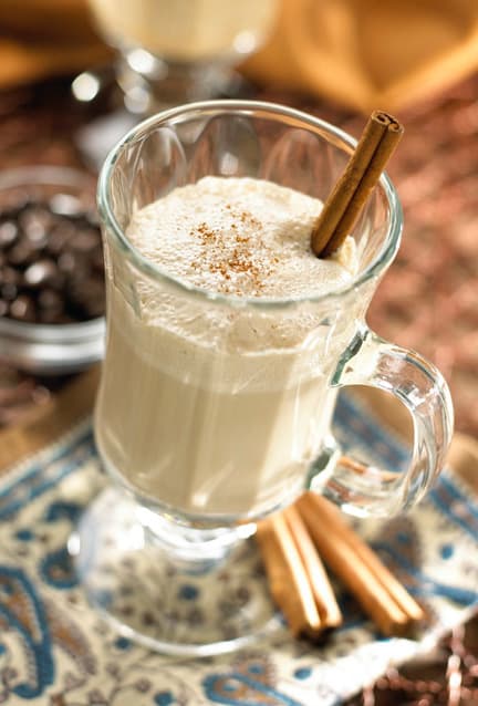 A glass mug filled with a creamy beverage, topped with ground spice and garnished with a cinnamon stick, sits on a patterned cloth alongside whole cinnamon sticks.