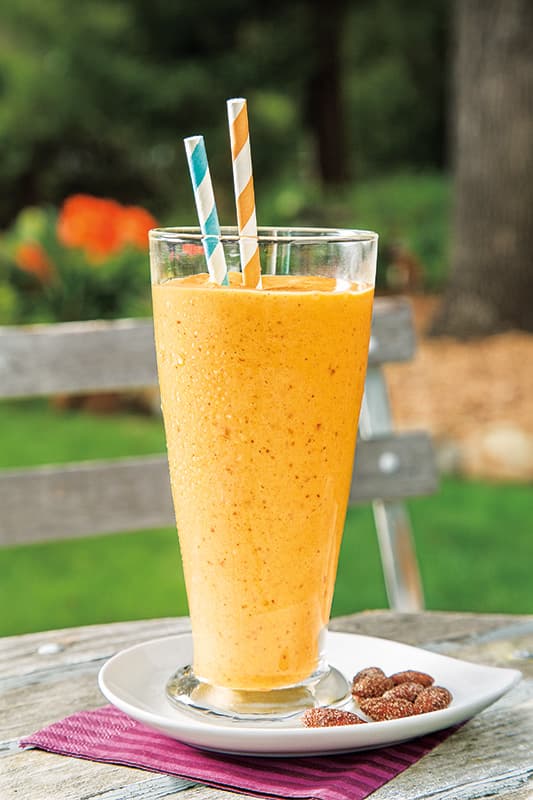 A tall glass of orange smoothie with two striped straws sits on a white plate beside small fruits, placed on an outdoor table with a green, blurred background.