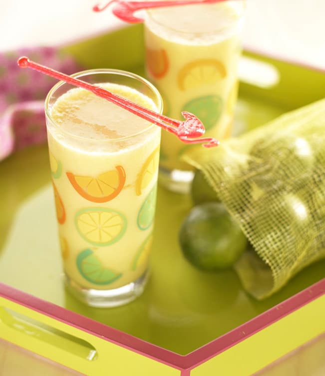 Two glasses of yellow juice with fruit slice designs and stir sticks sit on a green tray next to a mesh bag containing whole limes.