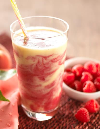 A glass of yellow and pink swirled smoothie with a straw, placed on a mat next to a bowl of fresh raspberries.