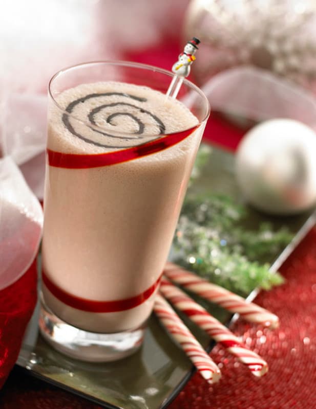 A glass of creamy beverage with chocolate swirl on top, red-striped pattern, a snowman stir stick, and three candy canes on the side, set on a holiday-themed table.
