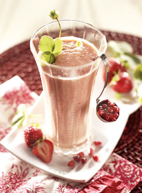 A glass of pink smoothie garnished with a green sprig, accompanied by strawberries and a spoon holding pomegranate seeds on a white plate.