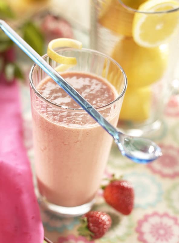A glass of strawberry smoothie with a blue spoon and lemon twist garnish, next to fresh strawberries and a glass jar of lemon slices.