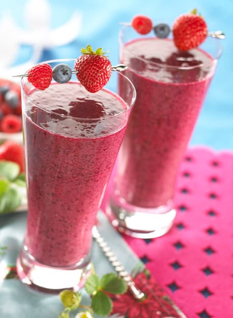 Two glasses of berry smoothie garnished with strawberries, raspberries, and blueberries on skewers, placed on a blue and pink surface with berries in the background.