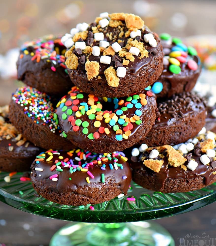 A stack of chocolate glazed donuts on a green glass plate, decorated with colorful sprinkles, mini marshmallows, and crushed cookies.