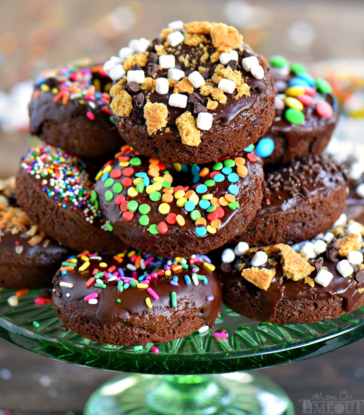 A stack of chocolate glazed donuts on a green glass plate, decorated with colorful sprinkles, mini marshmallows, and crushed cookies.