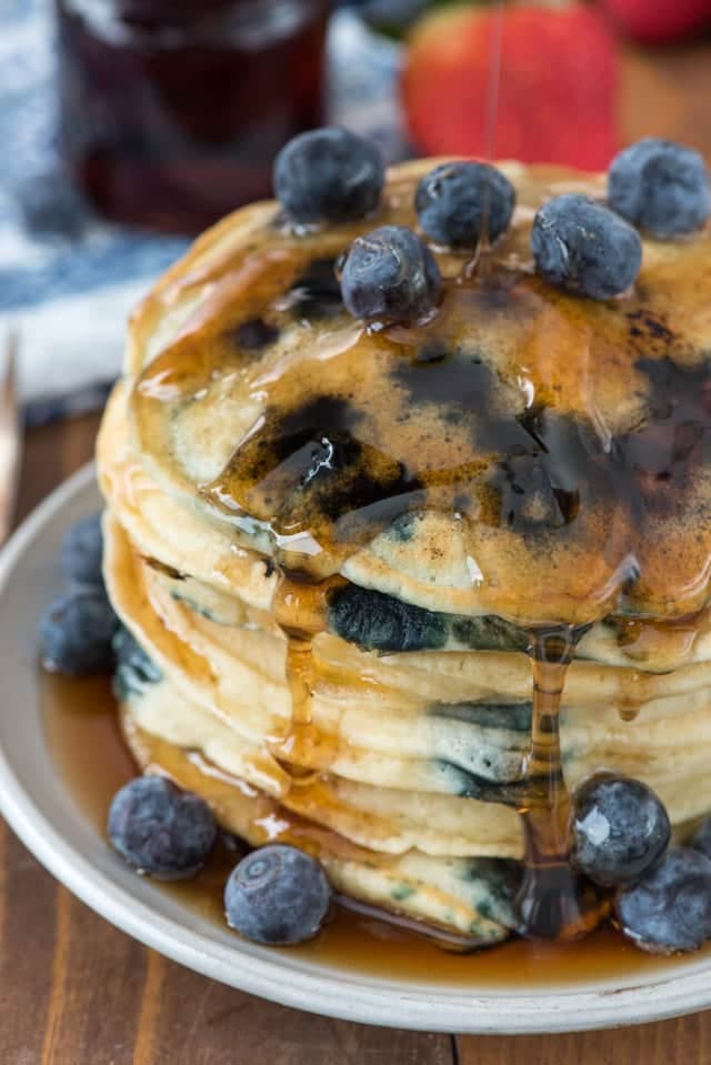 A stack of blueberry pancakes topped with fresh blueberries and drizzled with syrup sits on a plate.