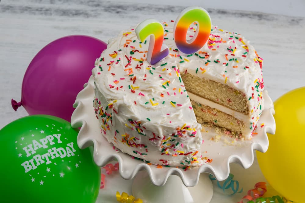 A birthday cake with rainbow 20 candles, white frosting, and sprinkles sits on a stand. A slice is missing. Green, purple, and yellow balloons are in the background.