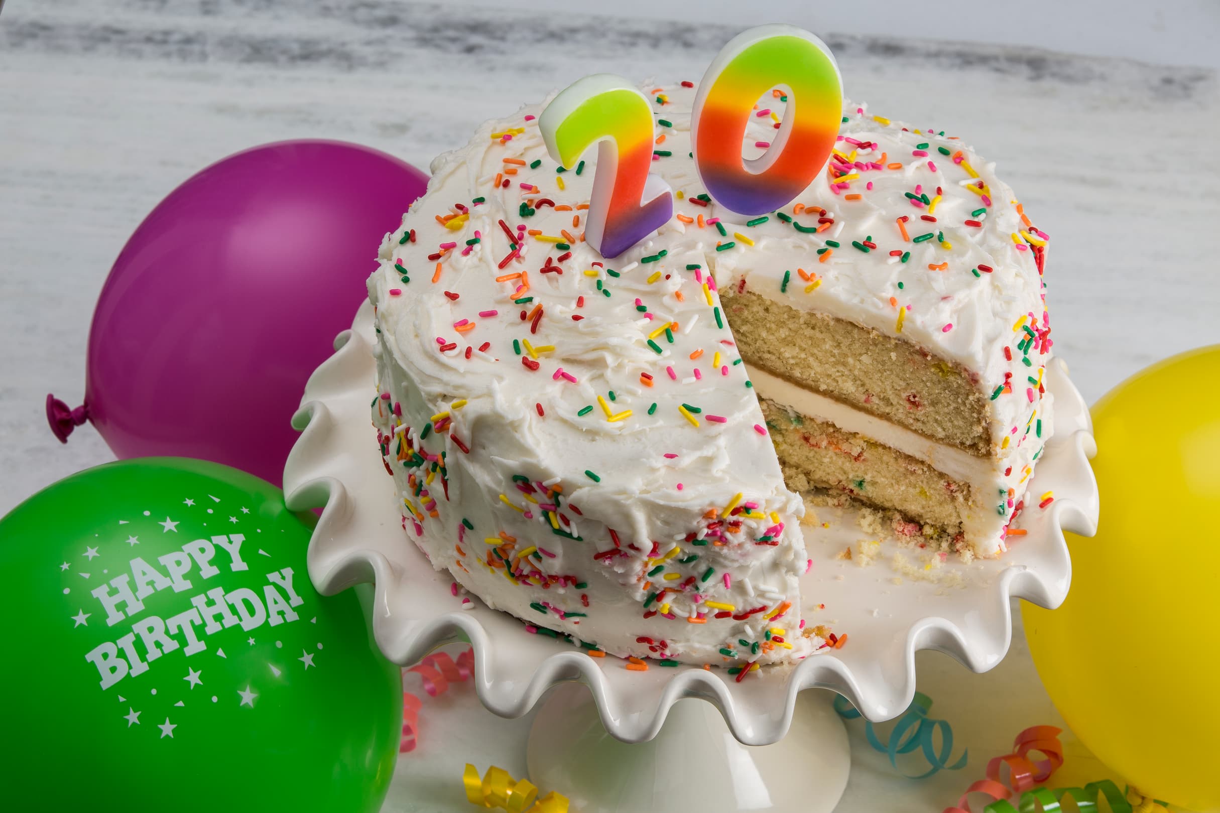 A birthday cake with rainbow 20 candles, white frosting, and sprinkles sits on a stand. A slice is missing. Green, purple, and yellow balloons are in the background.