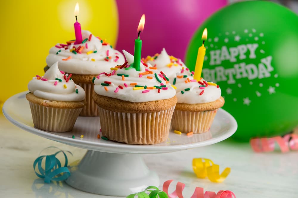 Four cupcakes with white frosting, sprinkles, and lit candles on a cake stand, surrounded by colorful balloons and streamers.