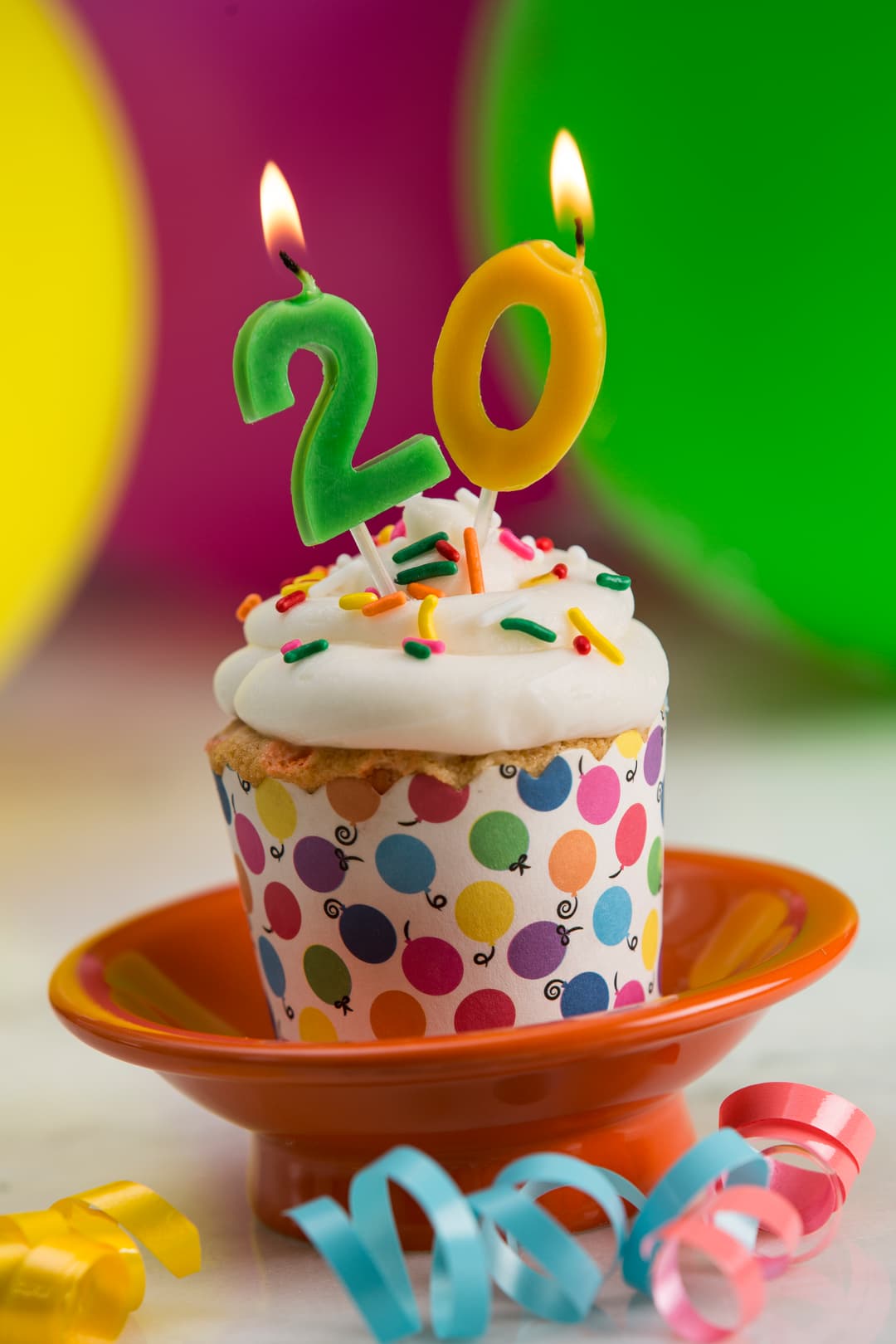 Cupcake with white icing, sprinkles, and lit candles shaped as the number 20, placed on an orange plate with colorful balloons and ribbons in the background.