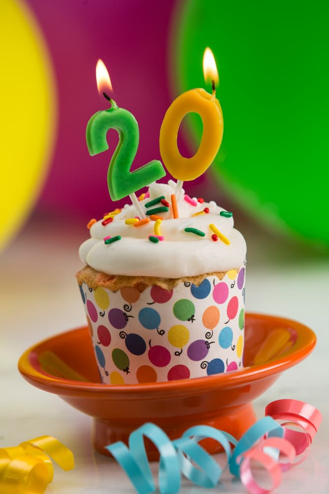 Cupcake with white icing, sprinkles, and lit candles shaped as the number 20, placed on an orange plate with colorful balloons and ribbons in the background.