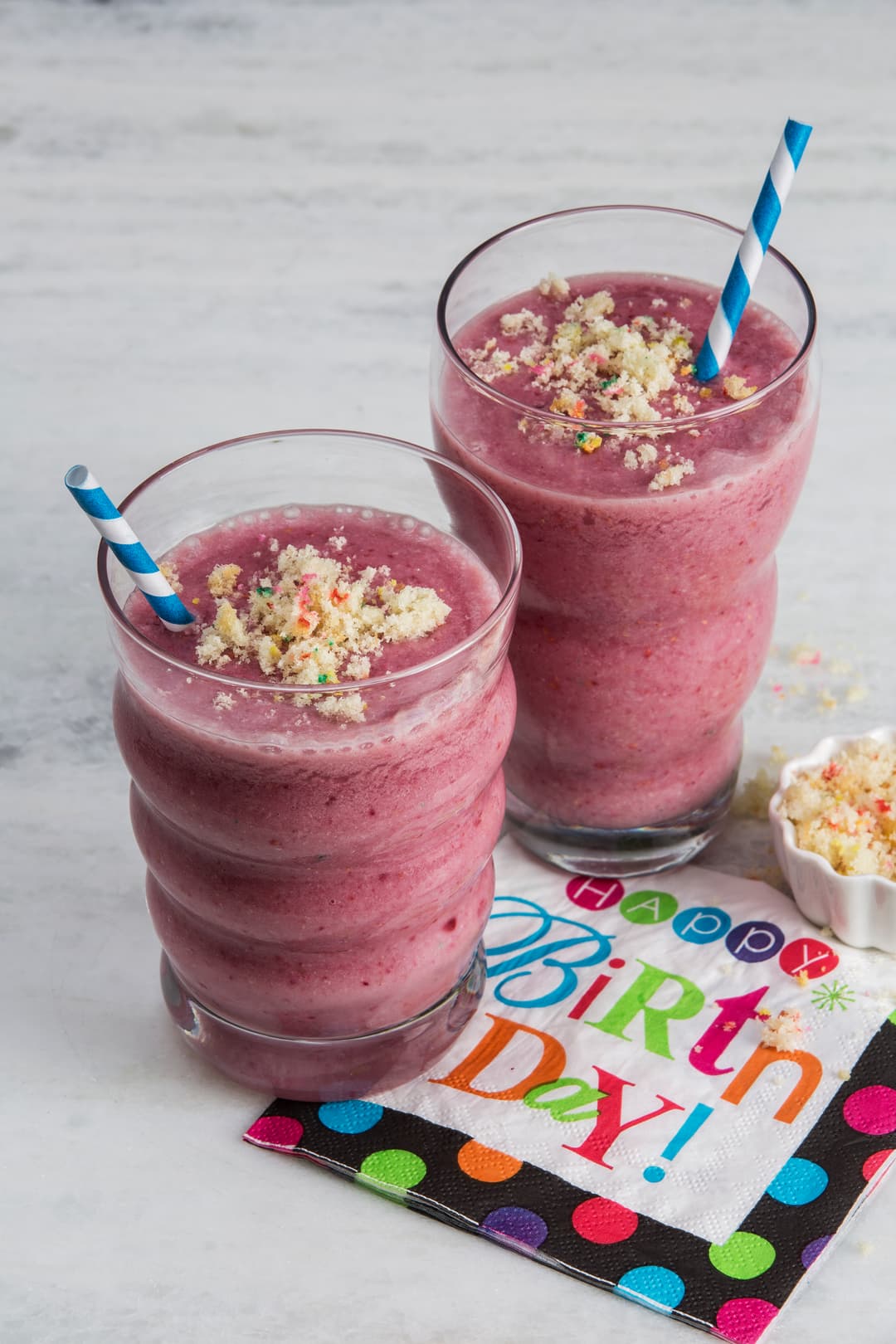 Two pink smoothies with blue striped straws are topped with crumbled cake, next to a colorful Happy Birthday! napkin on a light surface.