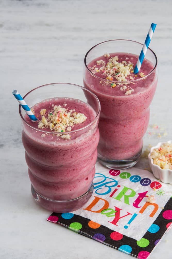 Two pink smoothies with blue striped straws are topped with crumbled cake, next to a colorful Happy Birthday! napkin on a light surface.