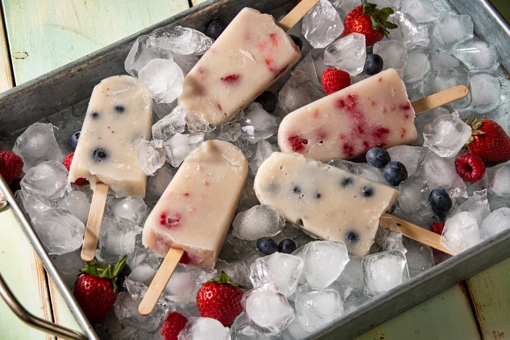 Five berry-filled popsicles are arranged on ice in a metal tray, surrounded by fresh strawberries and blueberries.
